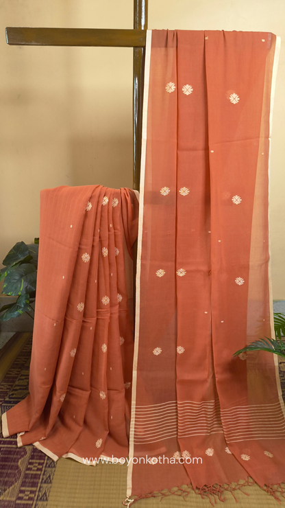Orangish peach buttery soft handloom cotton saree, with jamdani inspired handloom jacquard woven butas all over and braided tassels. Ideal for festive mornings 