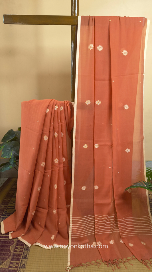 Orangish peach buttery soft handloom cotton saree, with jamdani inspired handloom jacquard woven butas all over and braided tassels. Ideal for festive mornings 