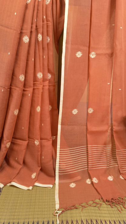 Orangish peach buttery soft handloom cotton saree, with jamdani inspired handloom jacquard woven butas all over and braided tassels. Ideal for festive mornings 
