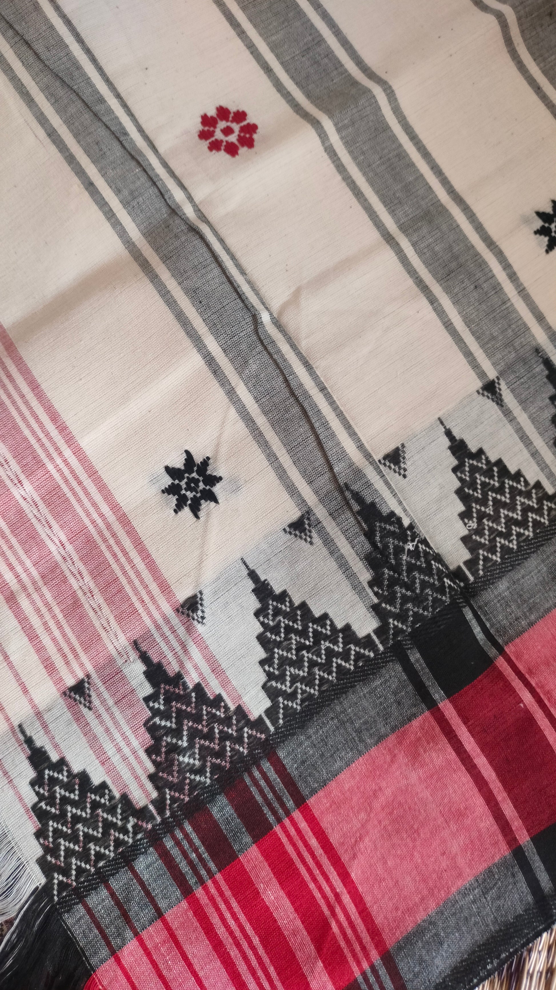 Close-up of Dhonekhali handloom saree from Bengal, featuring a pristine white body adorned with red and black woven butis throughout. The dual borders—deep red at the lower edge and bold black at the upper edge—showcase intricate temple motifs, while the pallu ends with tassels. 