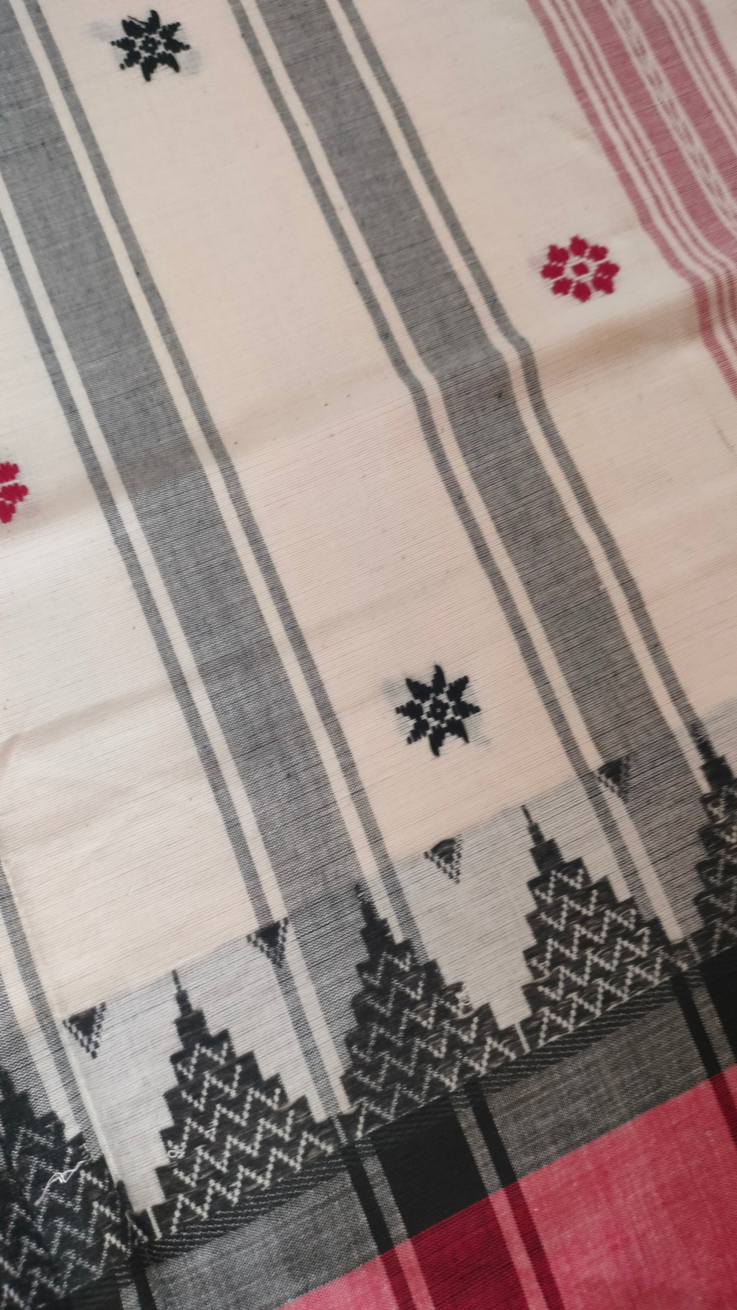 Dhonekhali handloom saree from Bengal, featuring a pristine white body adorned with red and black woven butis throughout. The dual borders—deep red at the lower edge and bold black at the upper edge—showcase intricate temple motifs, while the pallu ends with tassels. 