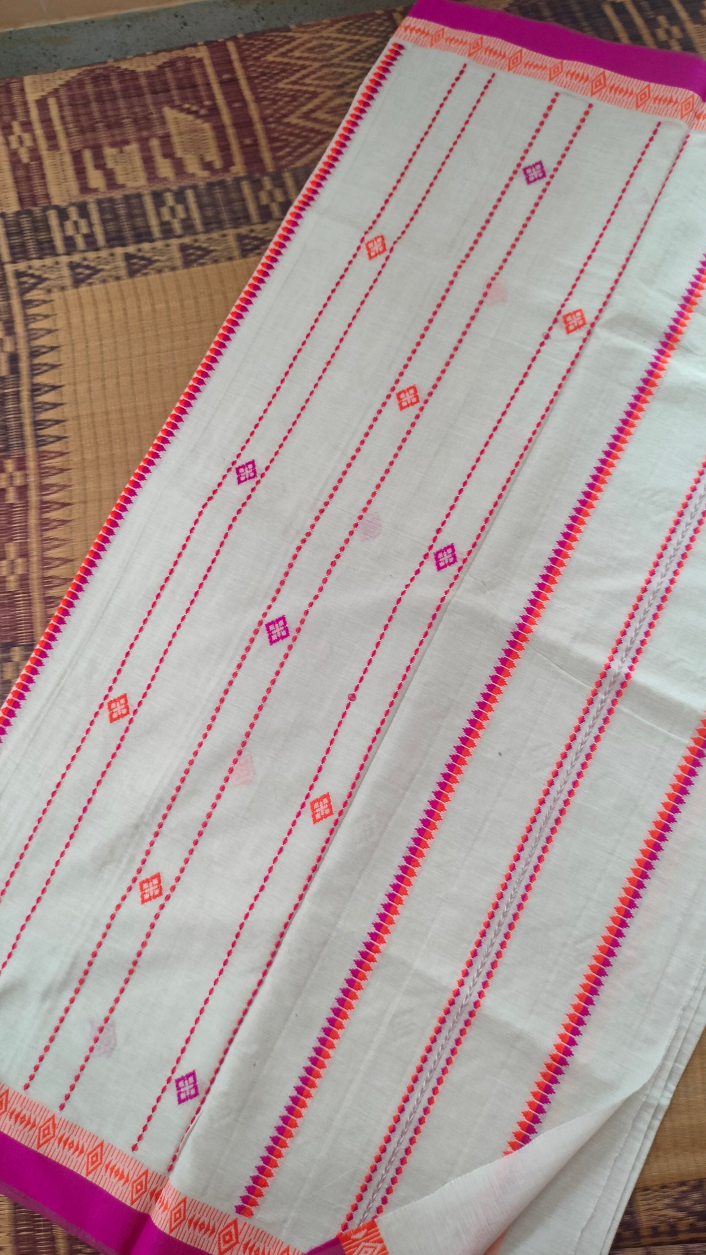 A white Dhonekhali handloom cotton saree with orange and pink woven motifs and bands on the saree. The border of the saree is pink with the inner edge having orange motifs.