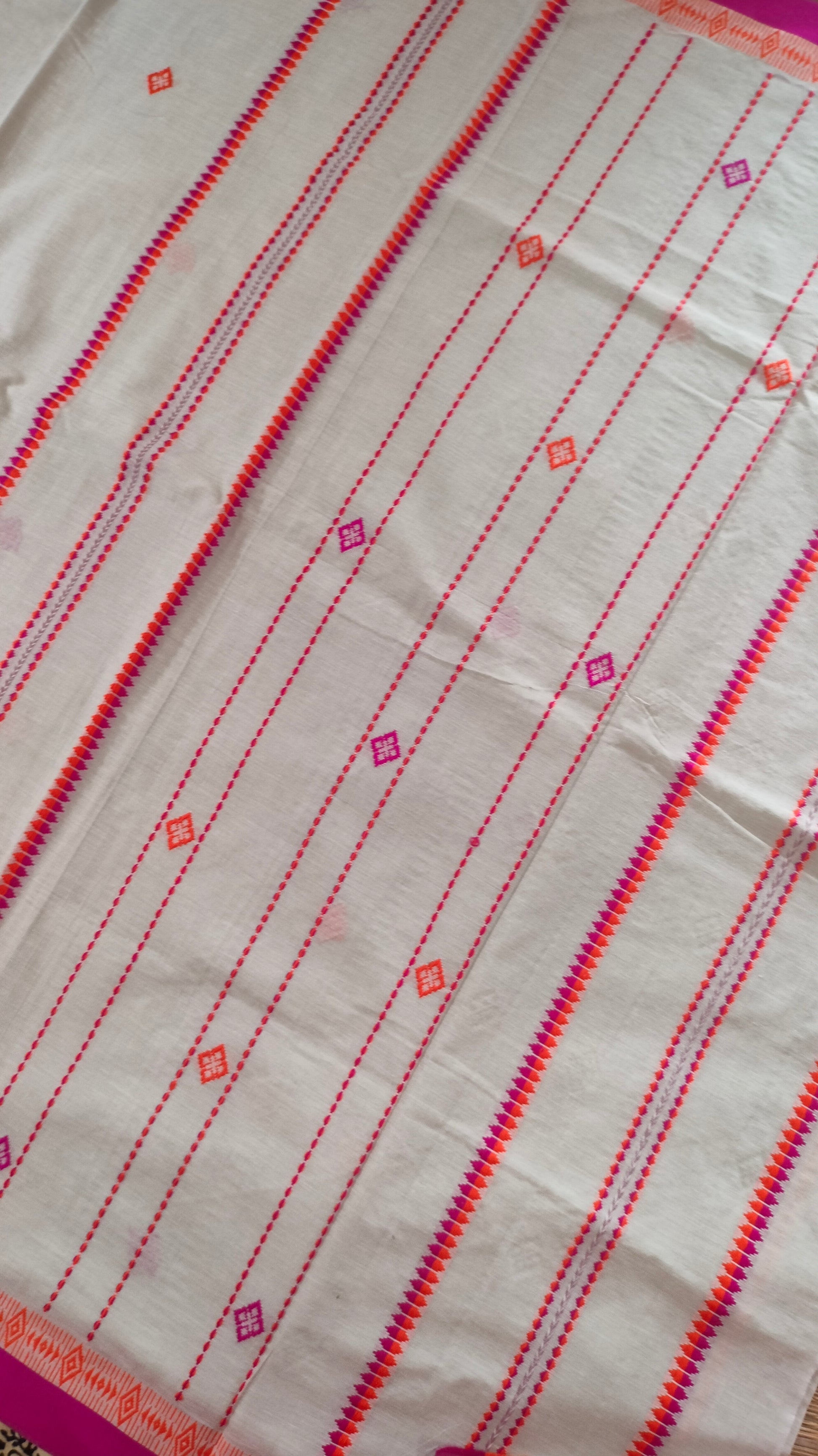 A white Dhonekhali handloom cotton saree with orange and pink woven motifs and bands on the saree. The border of the saree is pink with the inner edge having orange motifs.