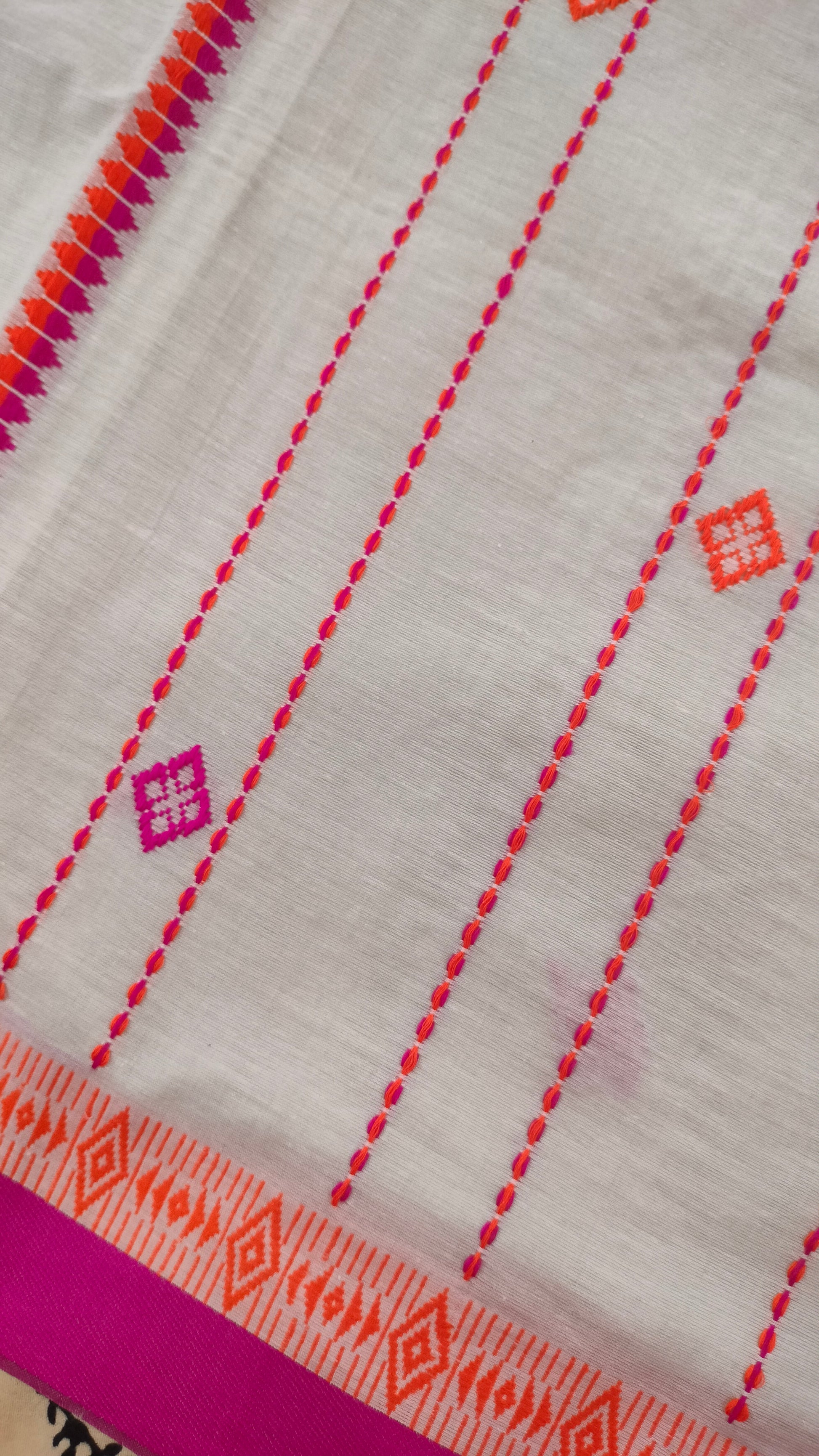 A white Dhonekhali handloom cotton saree with orange and pink woven motifs and bands on the saree. The border of the saree is pink with the inner edge having orange motifs.